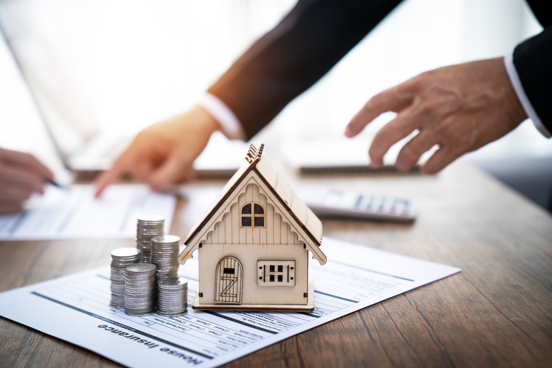 Wooden house model and stacked coins on a contract with two people discussing paperwork in a bright setting. Wooden house model and stacked coins on a contract with two people discussing paperwork in a bright setting.