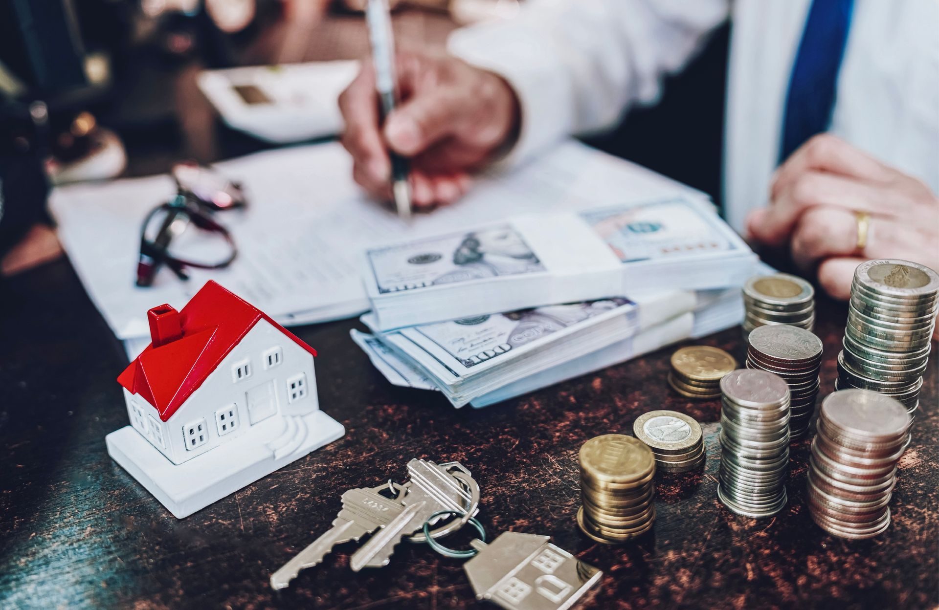 Person writing on documents near toy house, keys, money, and coins. Person writing on documents near toy house, keys, money, and coins.
