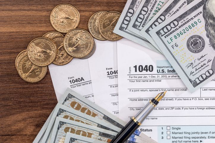 Tax forms, cash, and coins arranged on a wooden surface with a pen.