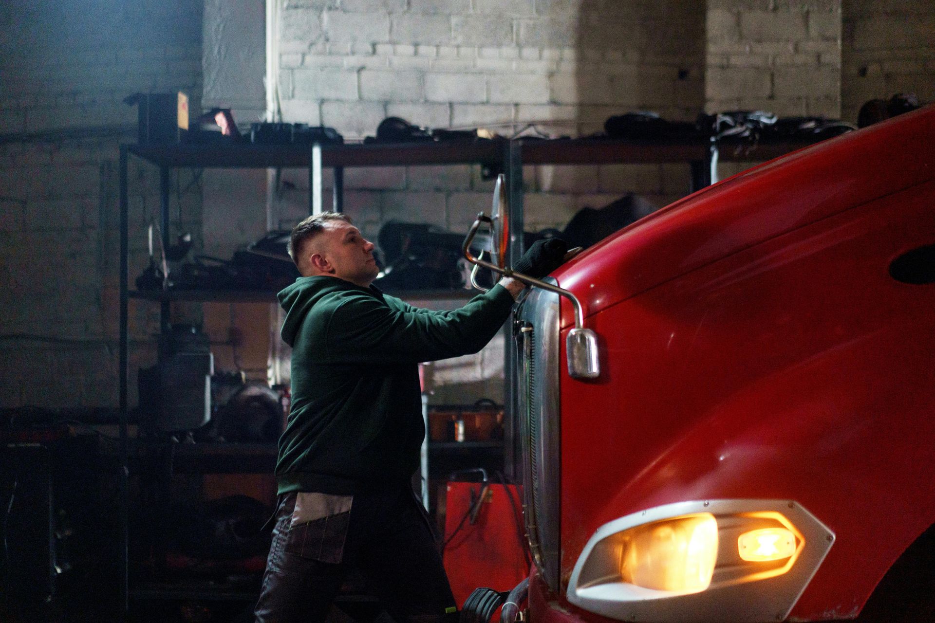 Man in green hoodie works on a red semi-truck in a dimly lit garage.