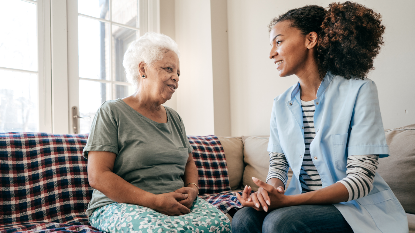 An elderly woman is sitting on a couch talking to a nurse.