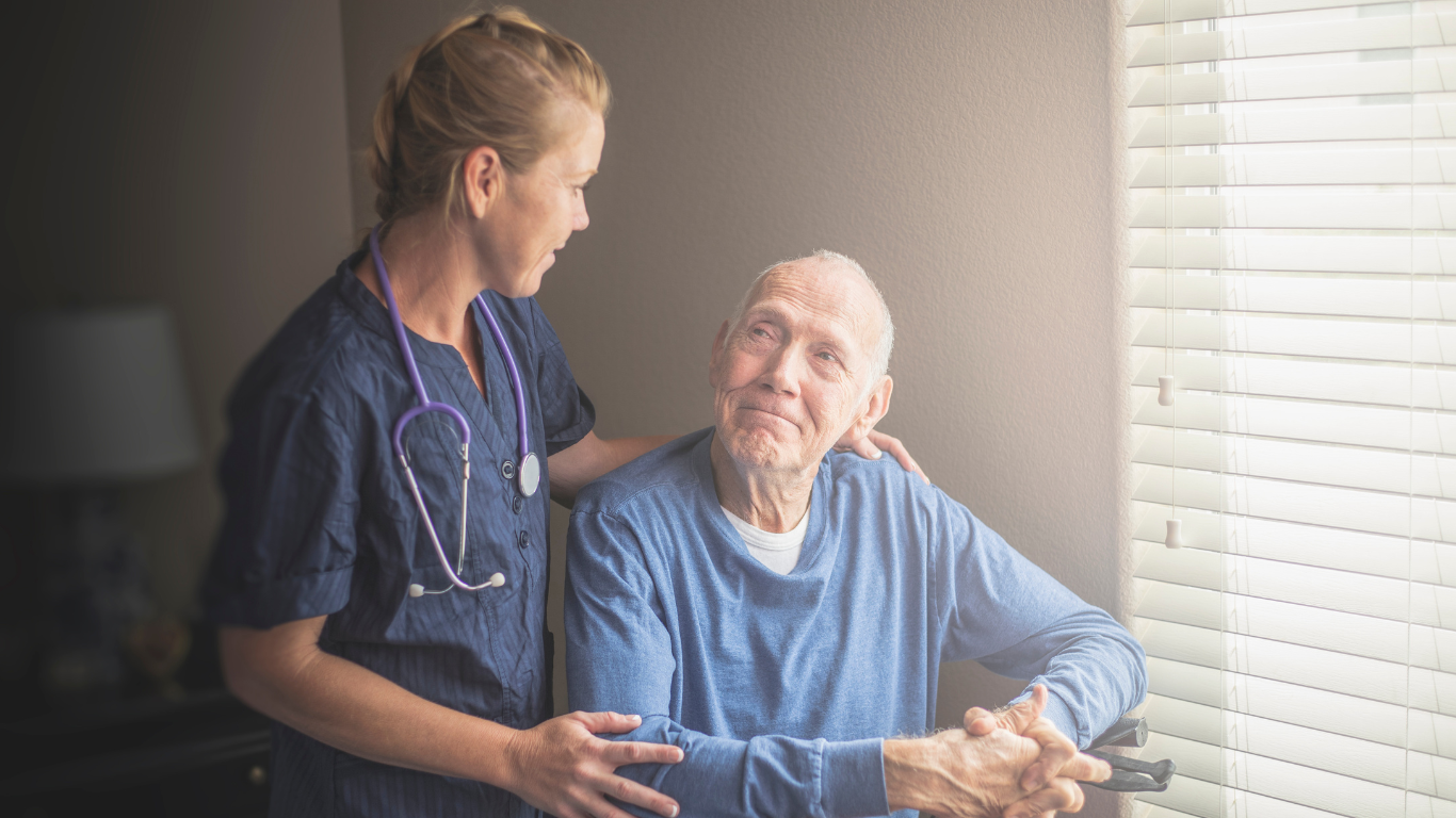 A nurse is standing next to an elderly man looking out of a window.