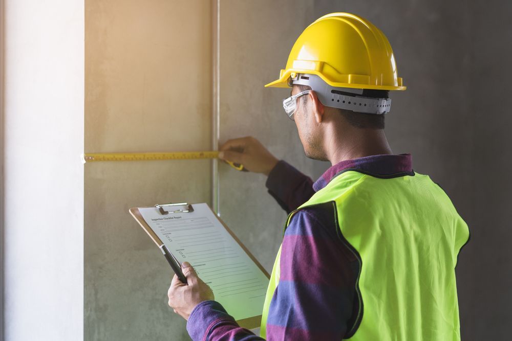 Construction worker in yellow hard hat measures and documents a wall, holding a clipboard.