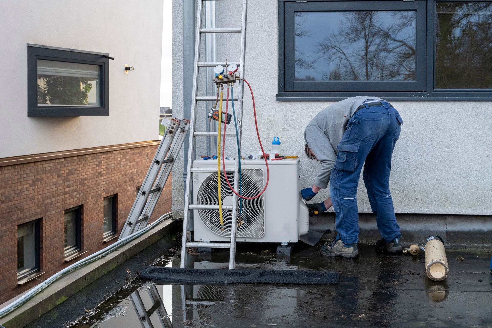 Person working on an AC unit on a rooftop, using a ladder and tools.