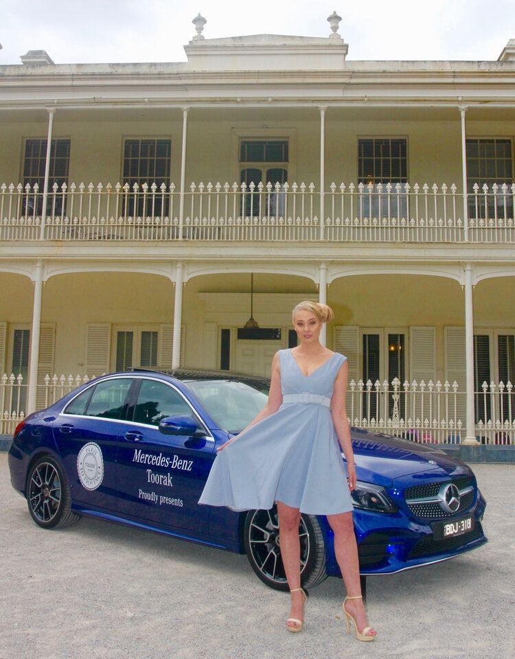 A woman in a light blue dress is standing next to a blue mercedes benz car.
