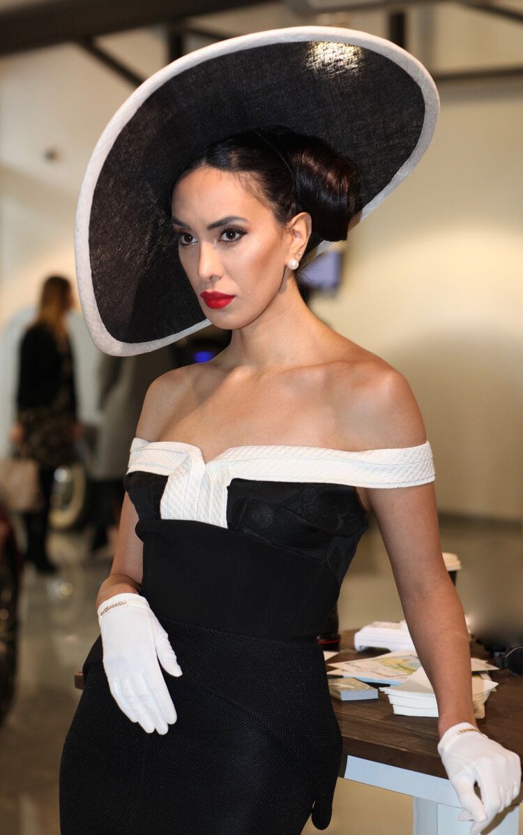 A woman in a black and white dress is wearing a large hat and white gloves.
