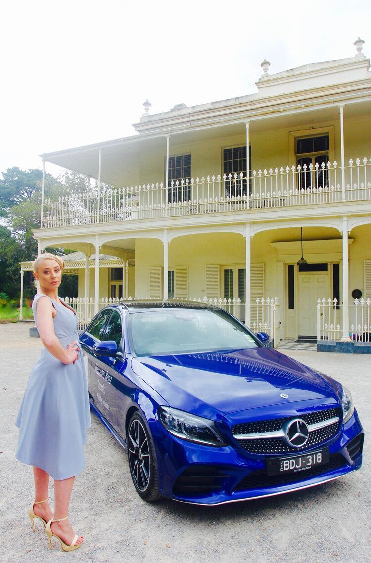 A woman in a blue dress is standing next to a blue car in front of a large white building.