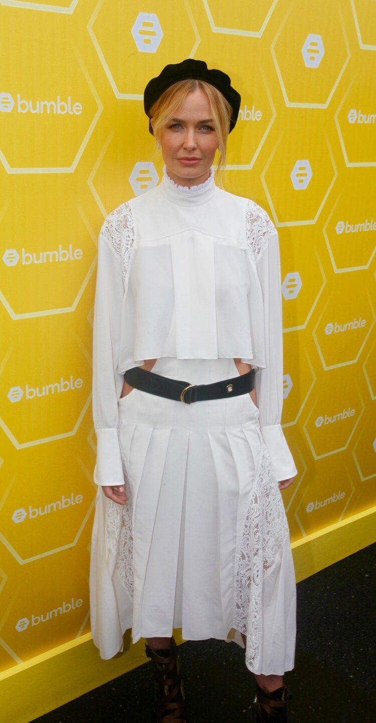 A woman in a white dress and black hat is standing in front of a yellow wall.