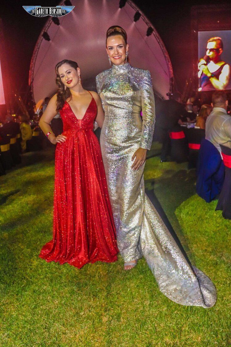 Two women in long dresses are standing next to each other on a lush green field.