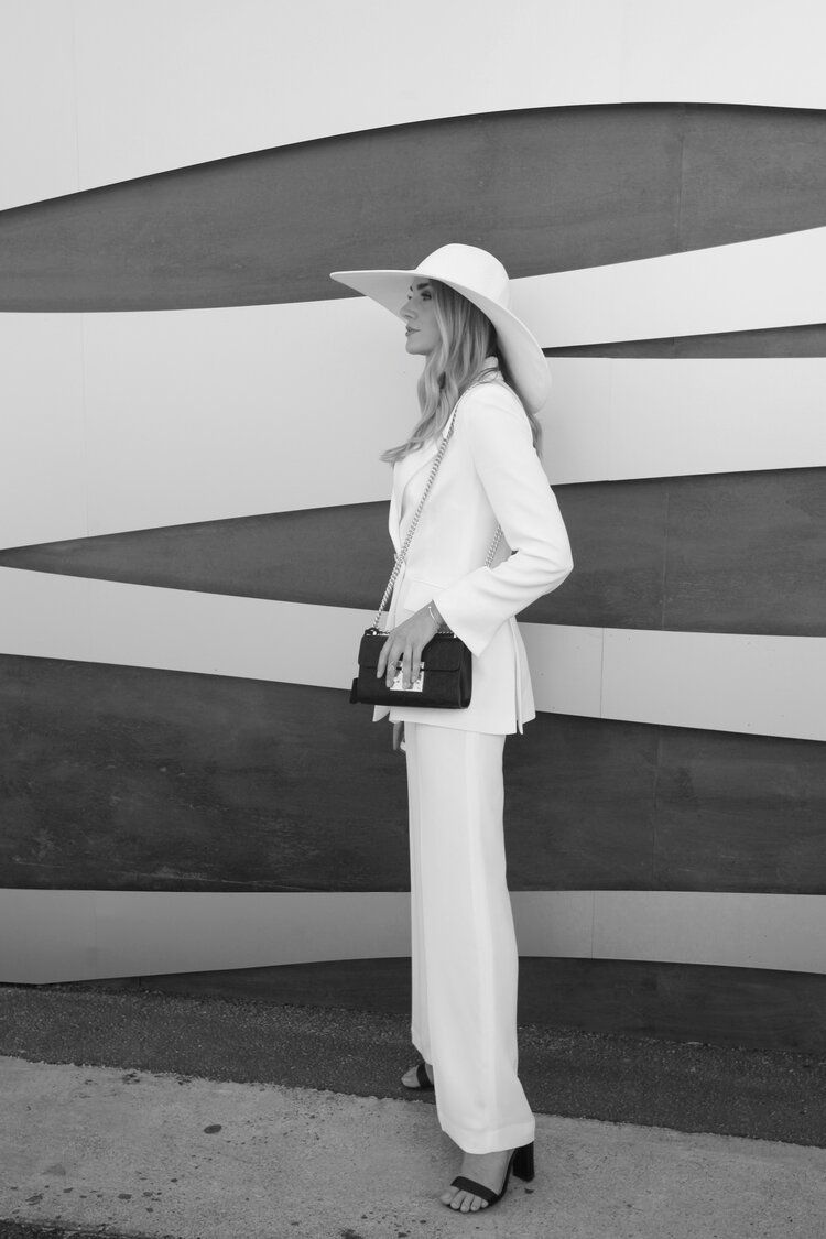 A woman in a white suit and hat is standing in front of a wall