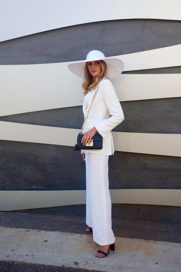 A woman in a white suit and hat is standing in front of a wall