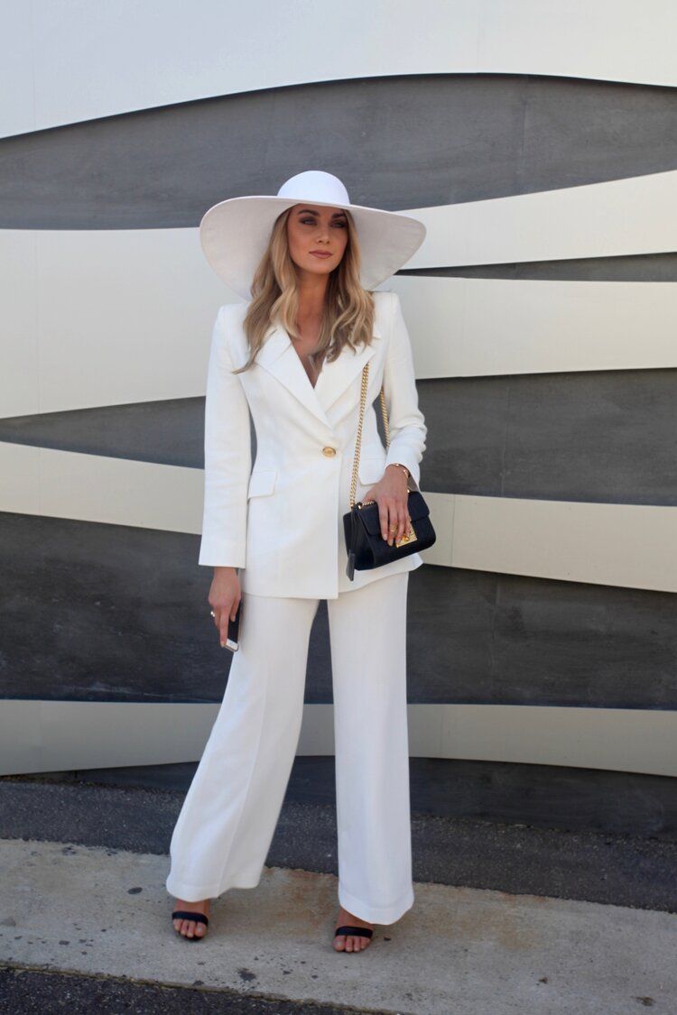 A woman in a white suit and hat is standing in front of a wall