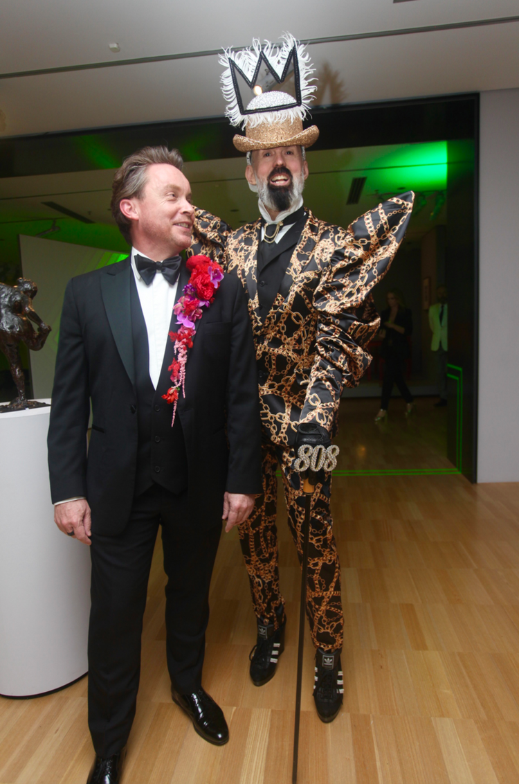 A man in a tuxedo stands next to a man in a costume
