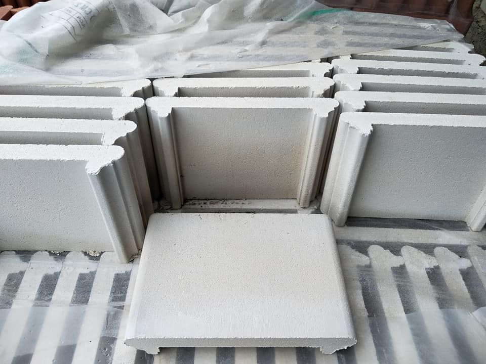 Pile di blocchi di cemento bianchi e quadrati con bordi decorativi, su una superficie a strisce, parzialmente coperti da un telo bianco.