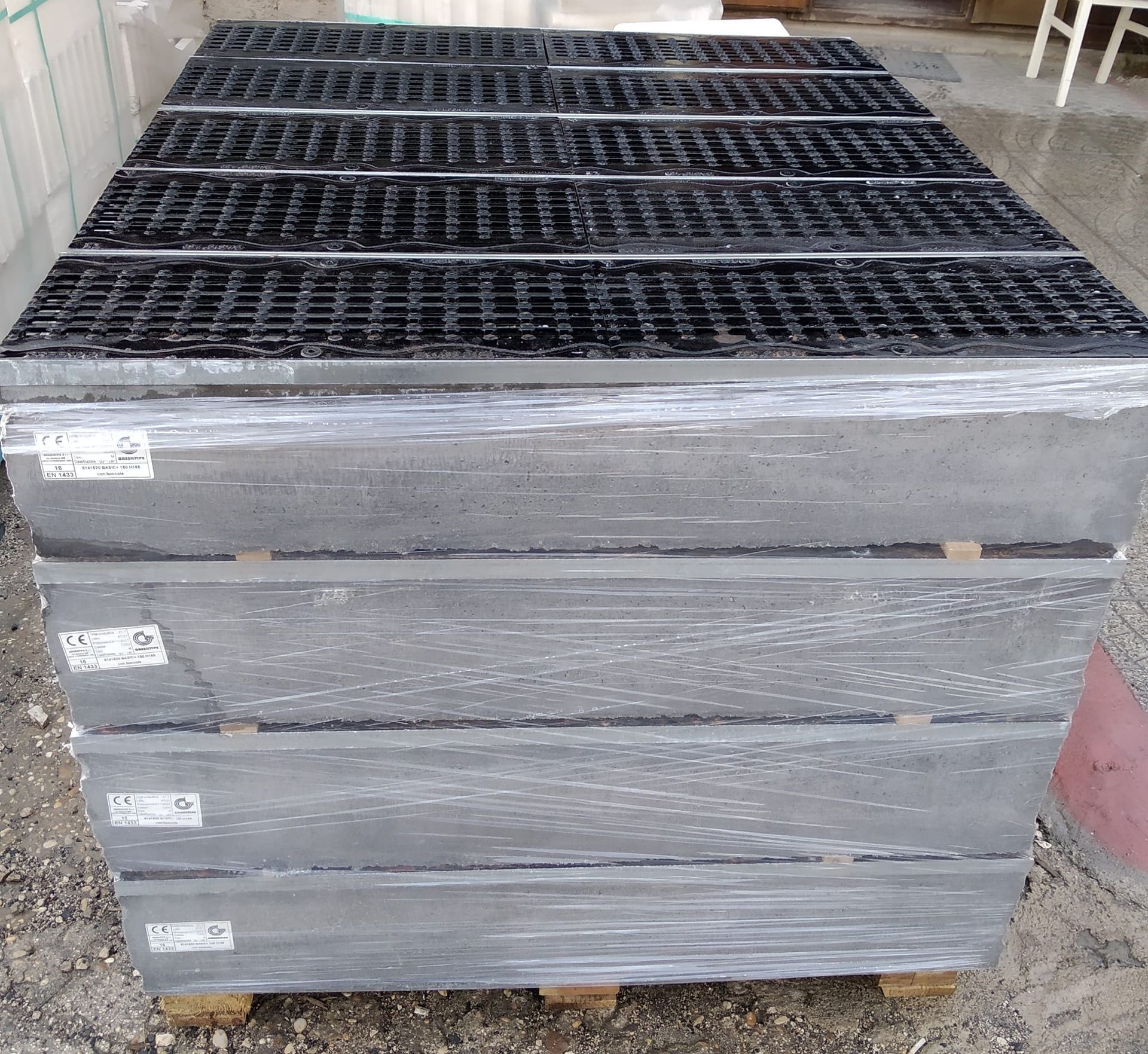 Pallet di basi di drenaggio in cemento grigio impilate con griglie metalliche nere sulla parte superiore, avvolte nella plastica.