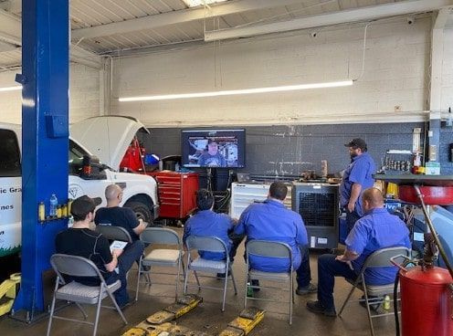 Technicians seated in a repair bay attentively watch a virtual training session on a large screen, featuring Diag Nation’s Adam Robertson and/or Eric Ziegler. The 2022 session took place at the shop, with tools, equipment, and a service vehicle visible in the background.