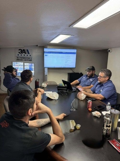 A group of team members sits around a conference table with food and drinks, engaged in a Lunch & Learn training session led by Service Manager Edgar Norris, with a digital dashboard displayed on the screen at 3A Automotive & Diesel Repair.