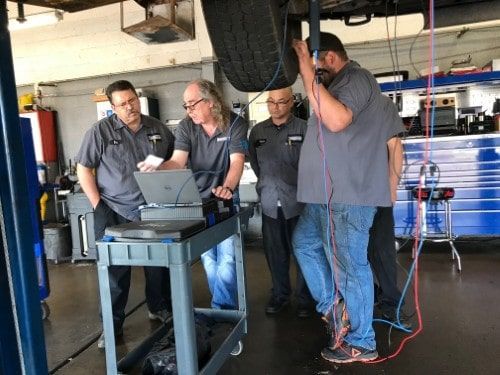 A group of automotive technicians gathered around a diagnostic laptop connected to a vehicle on a lift, receiving hands-on instruction from Eric Ziegler during Advanced Technical Training.