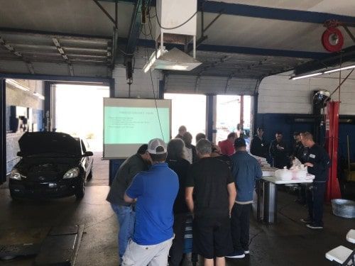 A training session in a garage setting, with a group of participants focused on a projected slide presentation. The session is part of Advanced Technical Training led by Eric Ziegler.