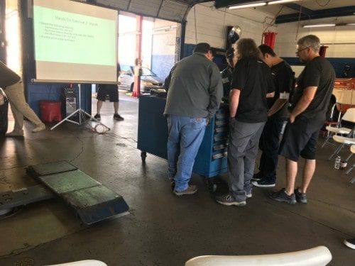 Several automotive professionals collaborate around a tool cart during a practical learning segment of the Advanced Technical Training with Eric Ziegler, while a slide presentation continues in the background.