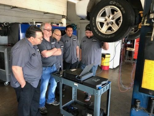 Technicians closely observe diagnostic data on a laptop connected to a raised vehicle, guided by Eric Ziegler as part of his Advanced Technical Training program.