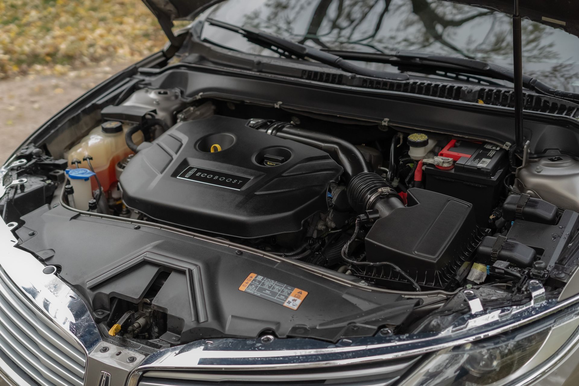 Car engine bay, black engine cover, battery, various tubes and containers, car hood open.