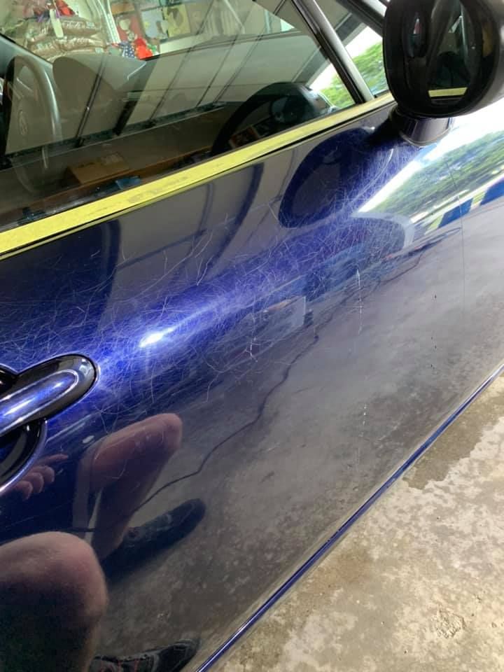 Blue car door covered in dust, with the reflection of a person taking the photo visible.