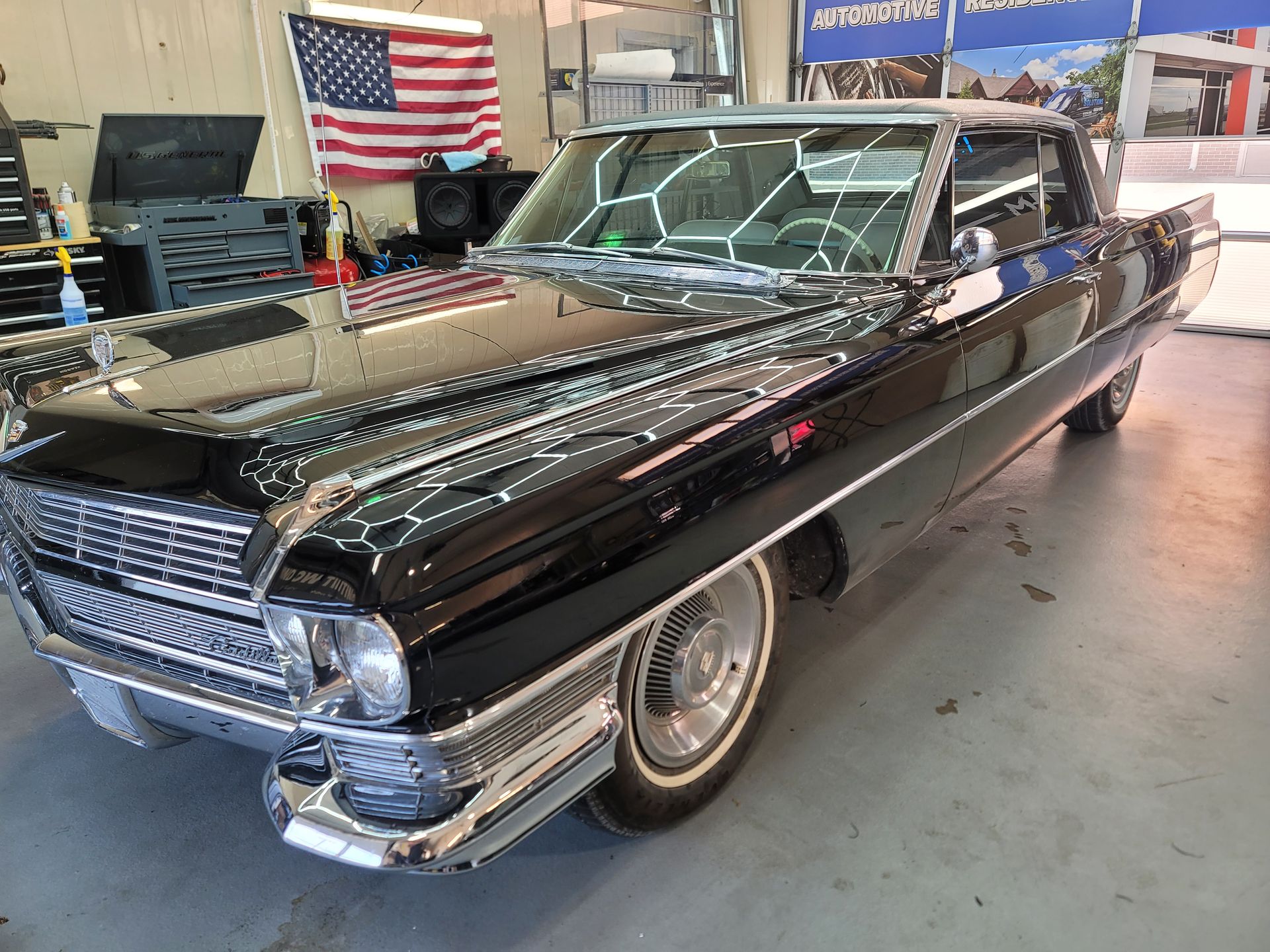 Black vintage Cadillac, chrome trim, white-wall tires, parked indoors, American flag in background.