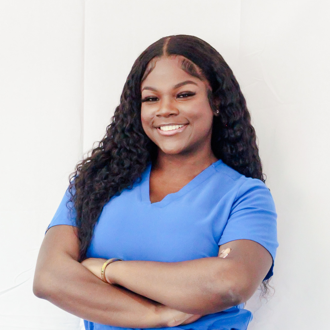 A woman in a blue scrub top is standing with her arms crossed and smiling.