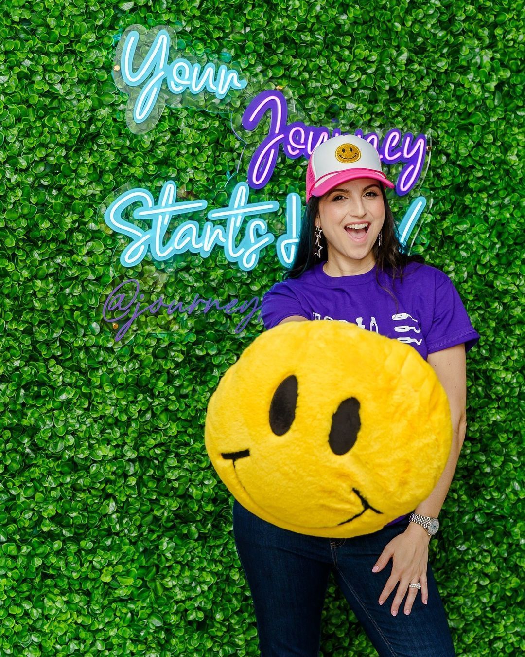 A woman in a purple shirt is holding a yellow smiley face pillow.