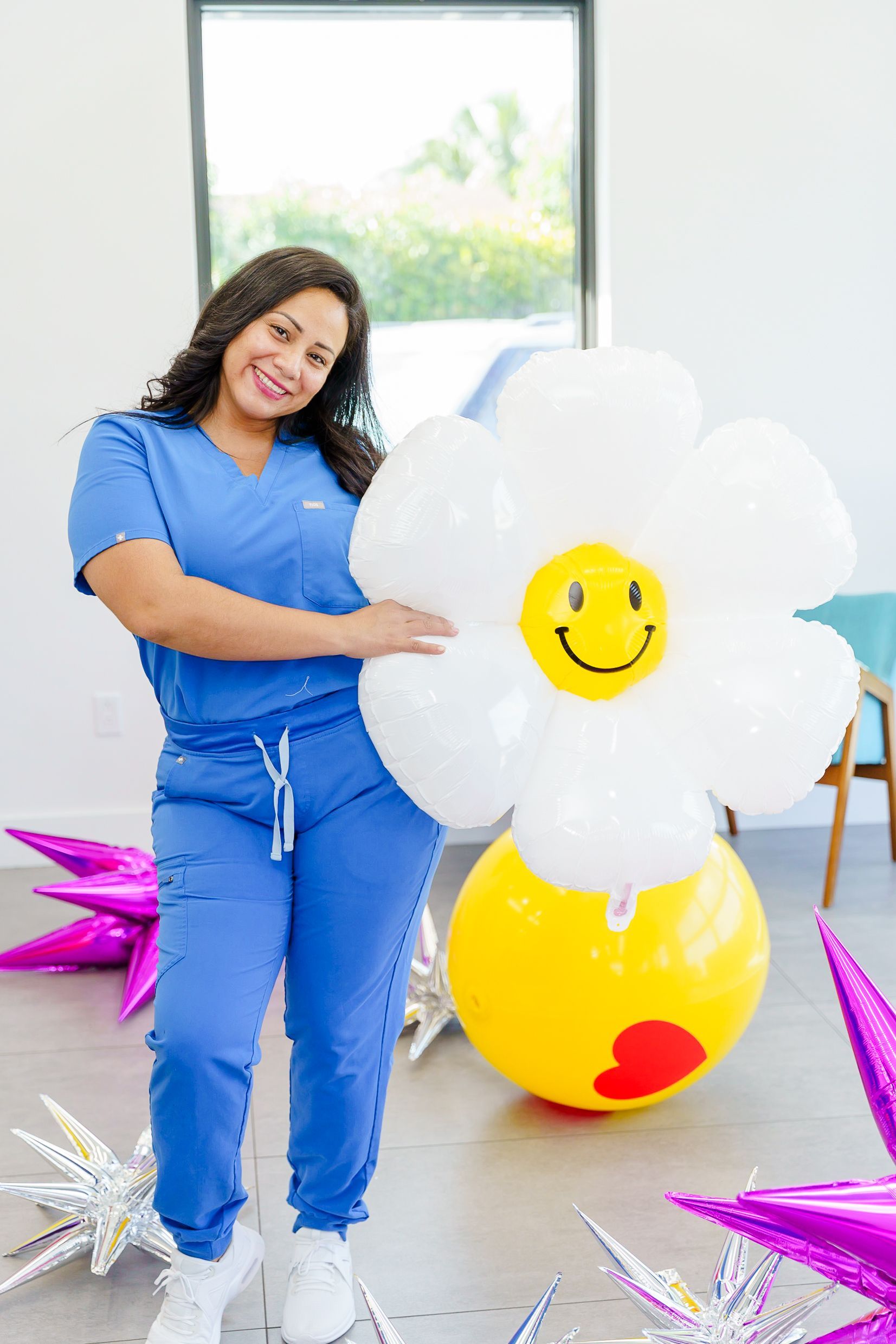 A woman in blue scrubs is holding balloons in the shape of a flower with a smiley face on it.