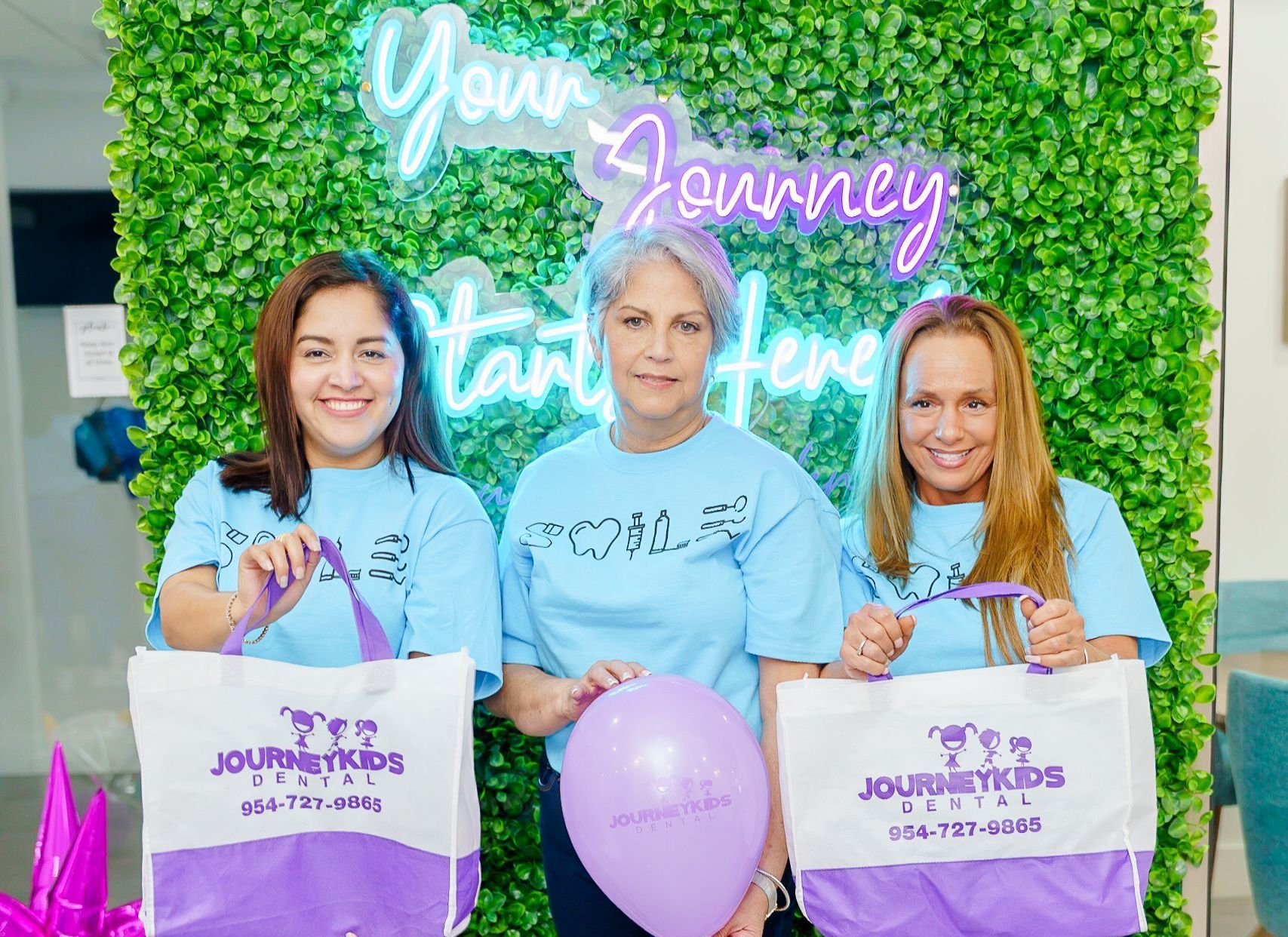 Three women are standing in front of a green wall holding balloons and bags.