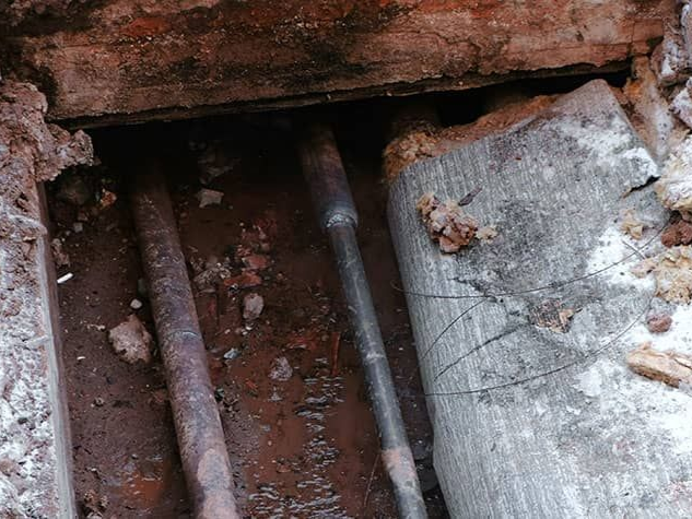 Pipes in a trench, partially covered. Brown dirt and a concrete slab are also visible.