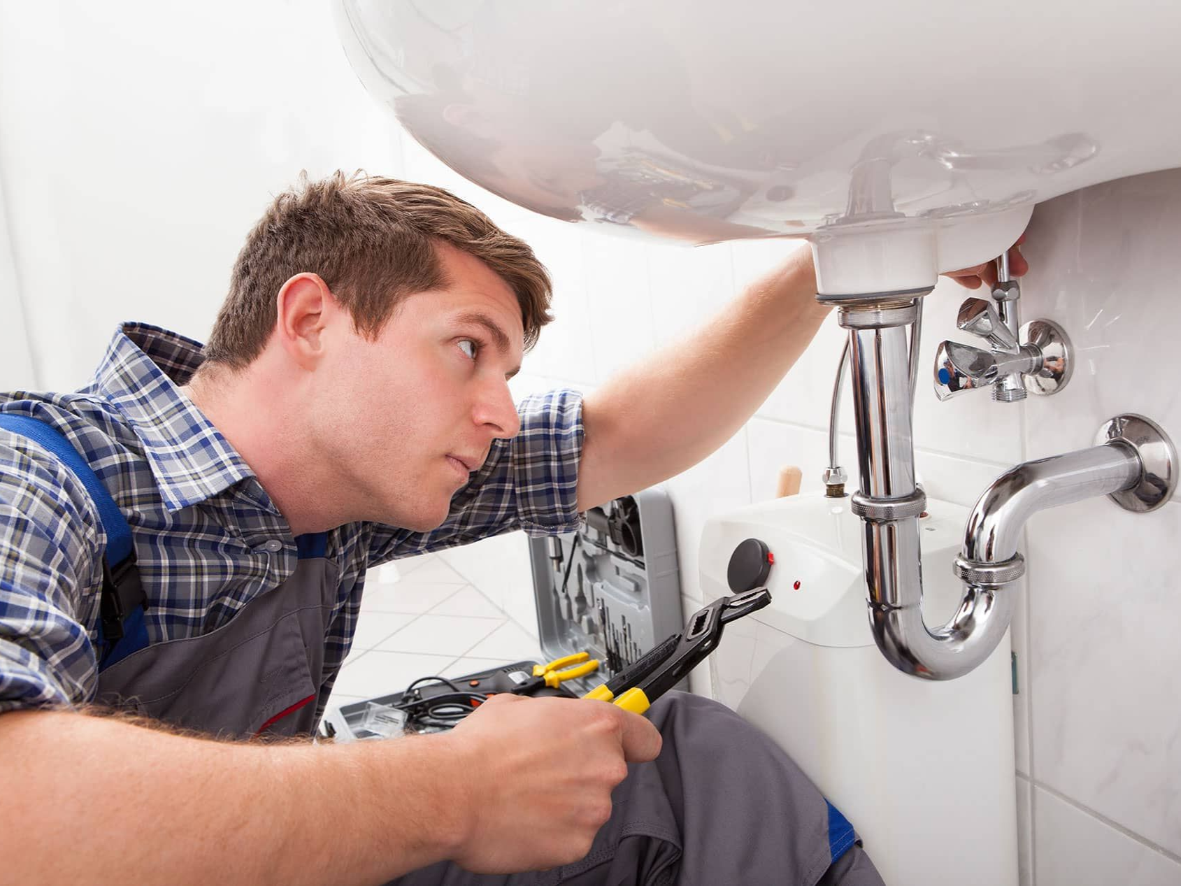 Plumber in blue and gray overalls working under a white sink with a wrench in a bathroom.