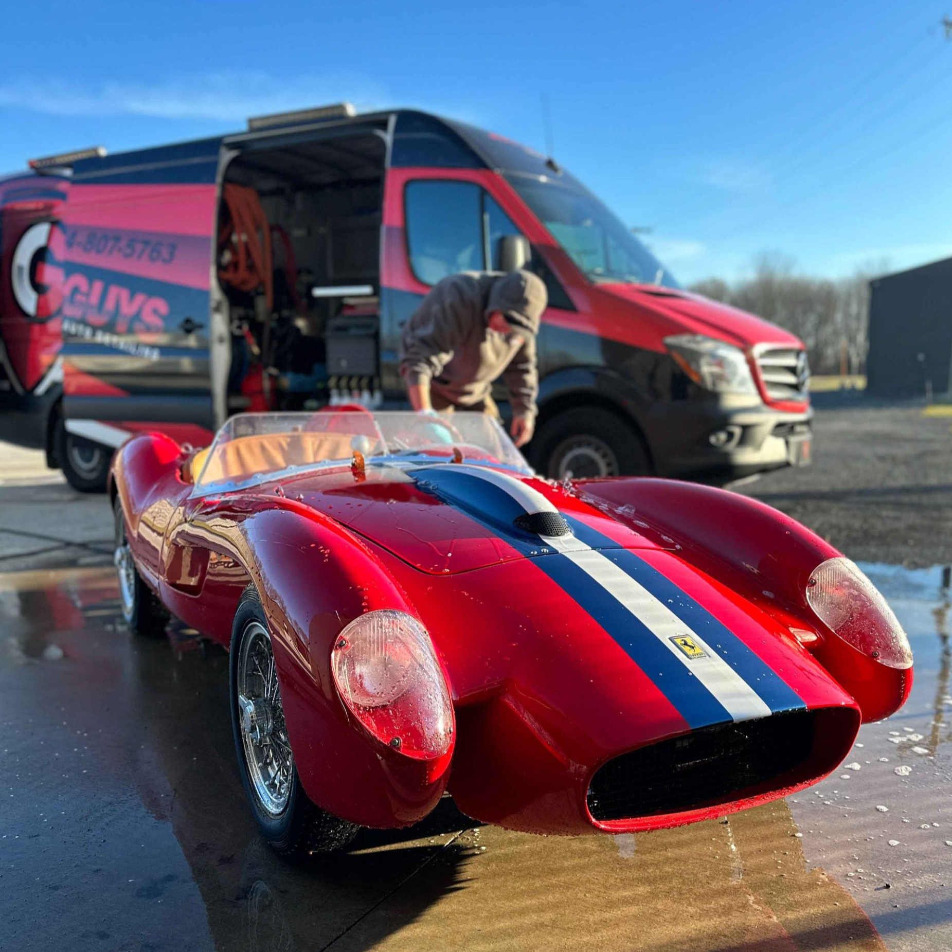 A red, white, and blue vintage-style sports car parked in front of a red service van with a person leaning inside.
