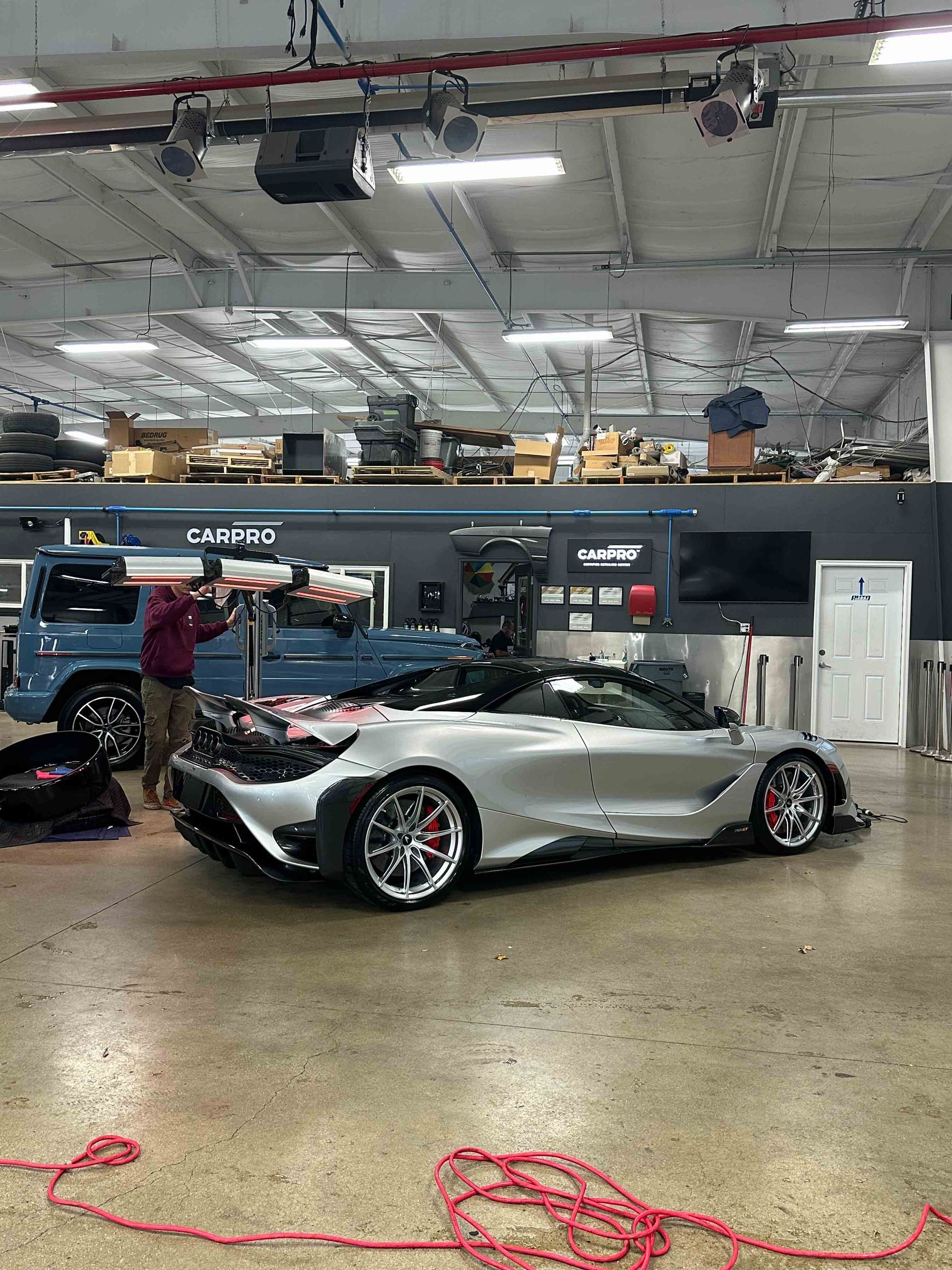 Silver sports car inside a workshop, surrounded by tools and equipment; a person is working on the car.