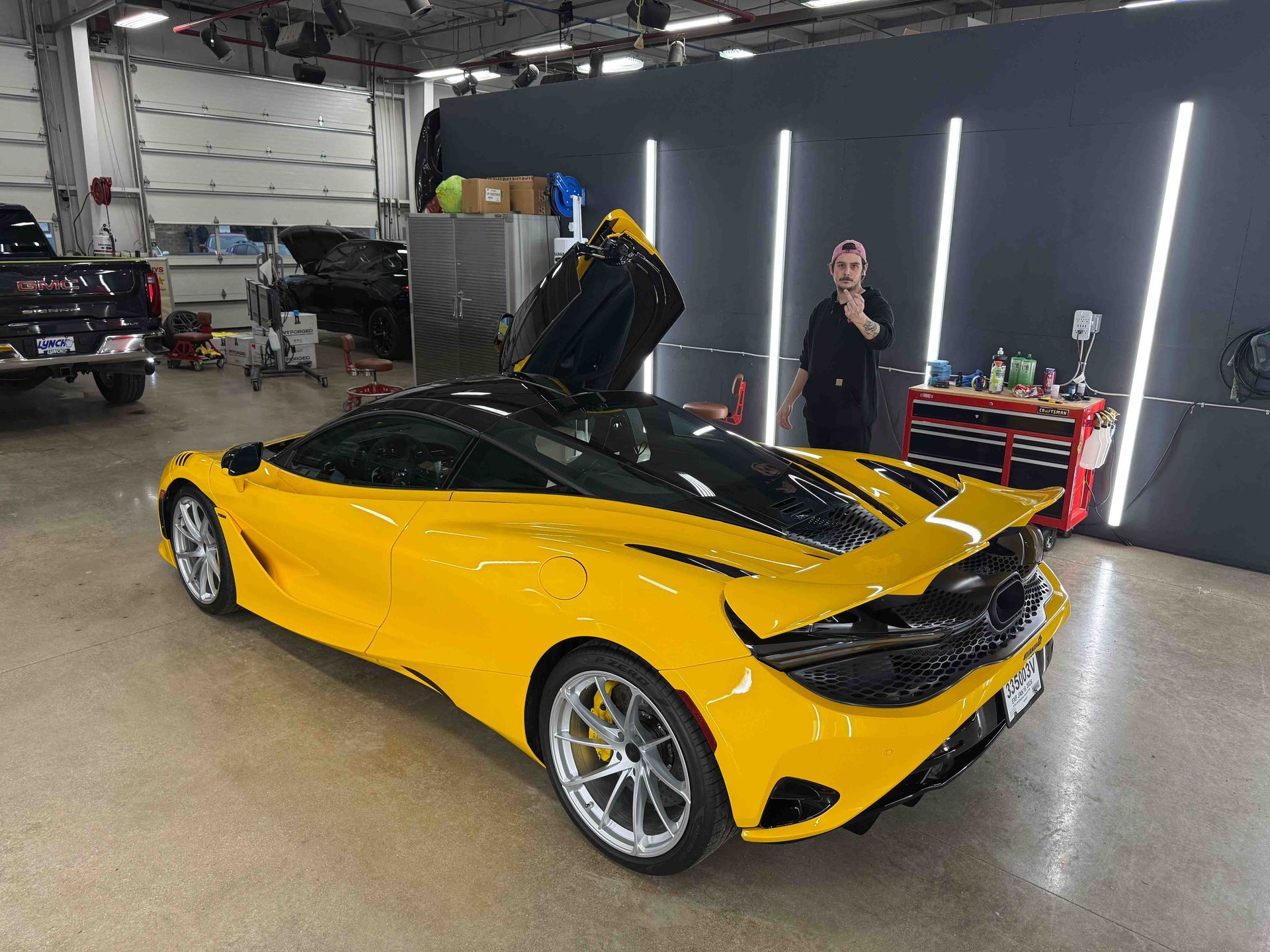 Yellow sports car with black top, doors open, in a garage with a person standing nearby.