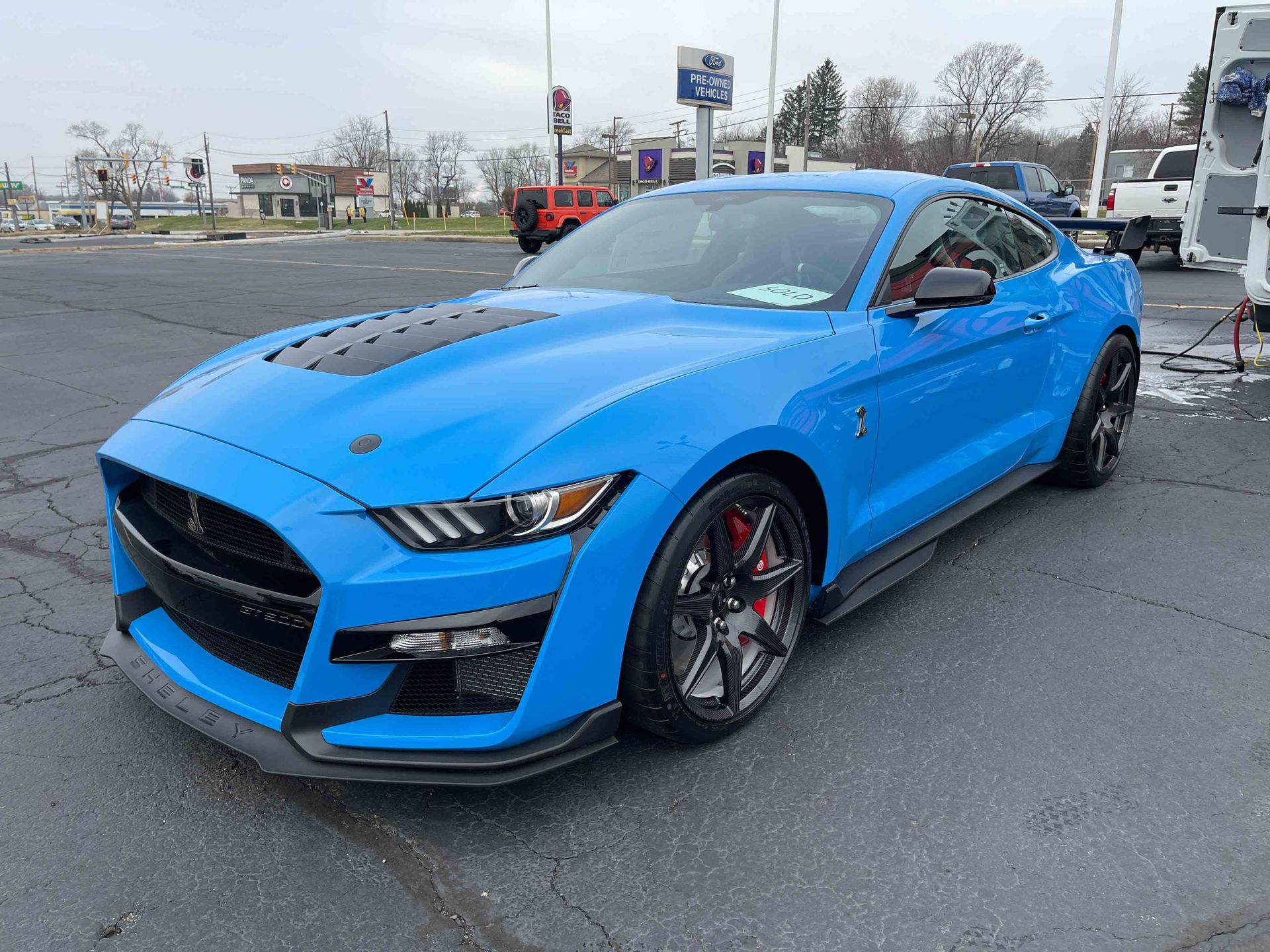 Blue Ford Shelby GT500 sports car with black racing stripes parked outside a dealership.