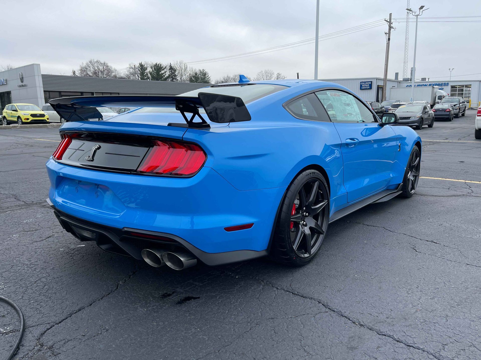 Blue Ford Mustang with a large rear wing parked outside a dealership on an overcast day.