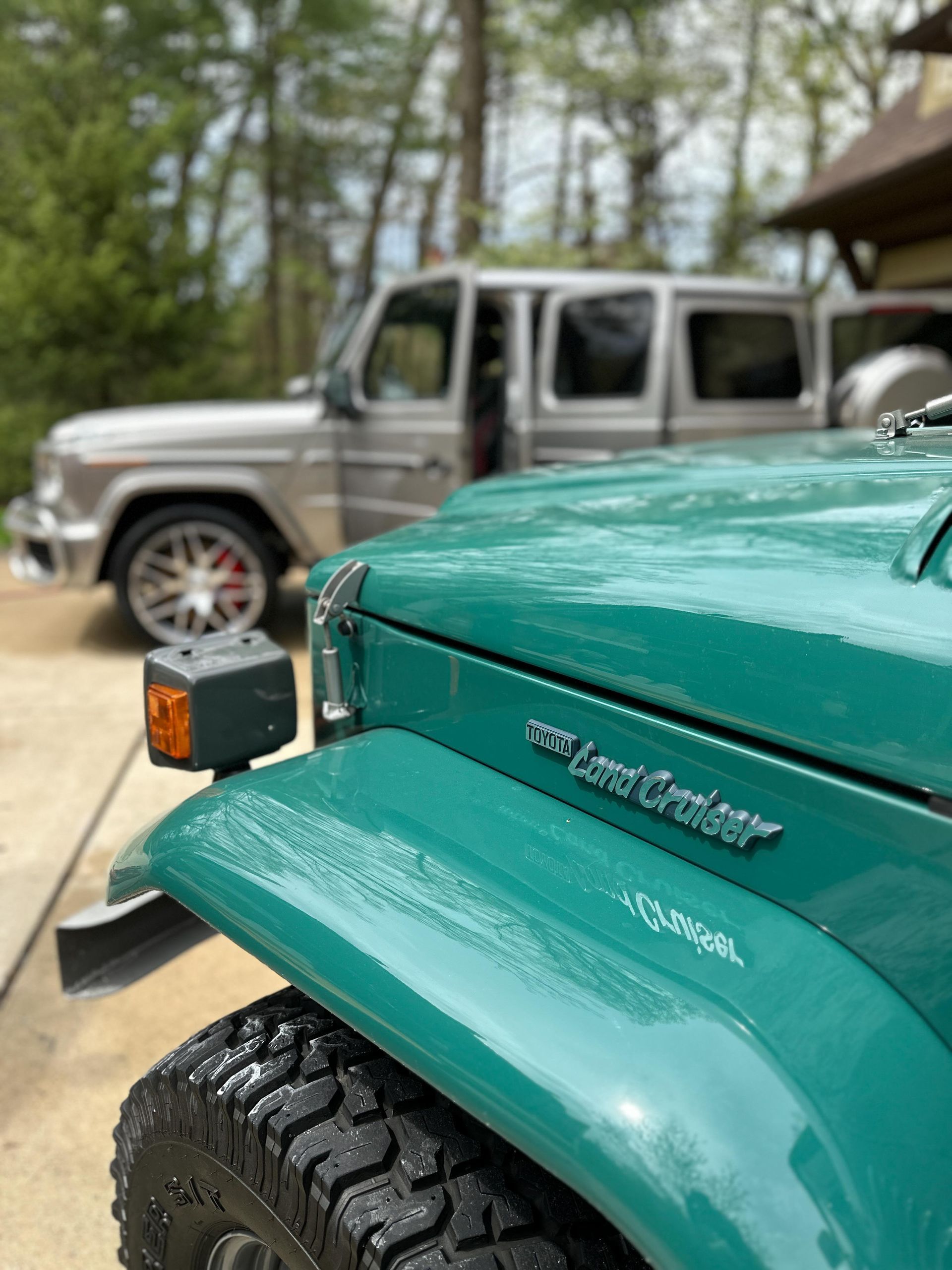 Teal vintage Toyota Land Cruiser with a silver Mercedes-Benz G-Class in the background. Outdoor setting.