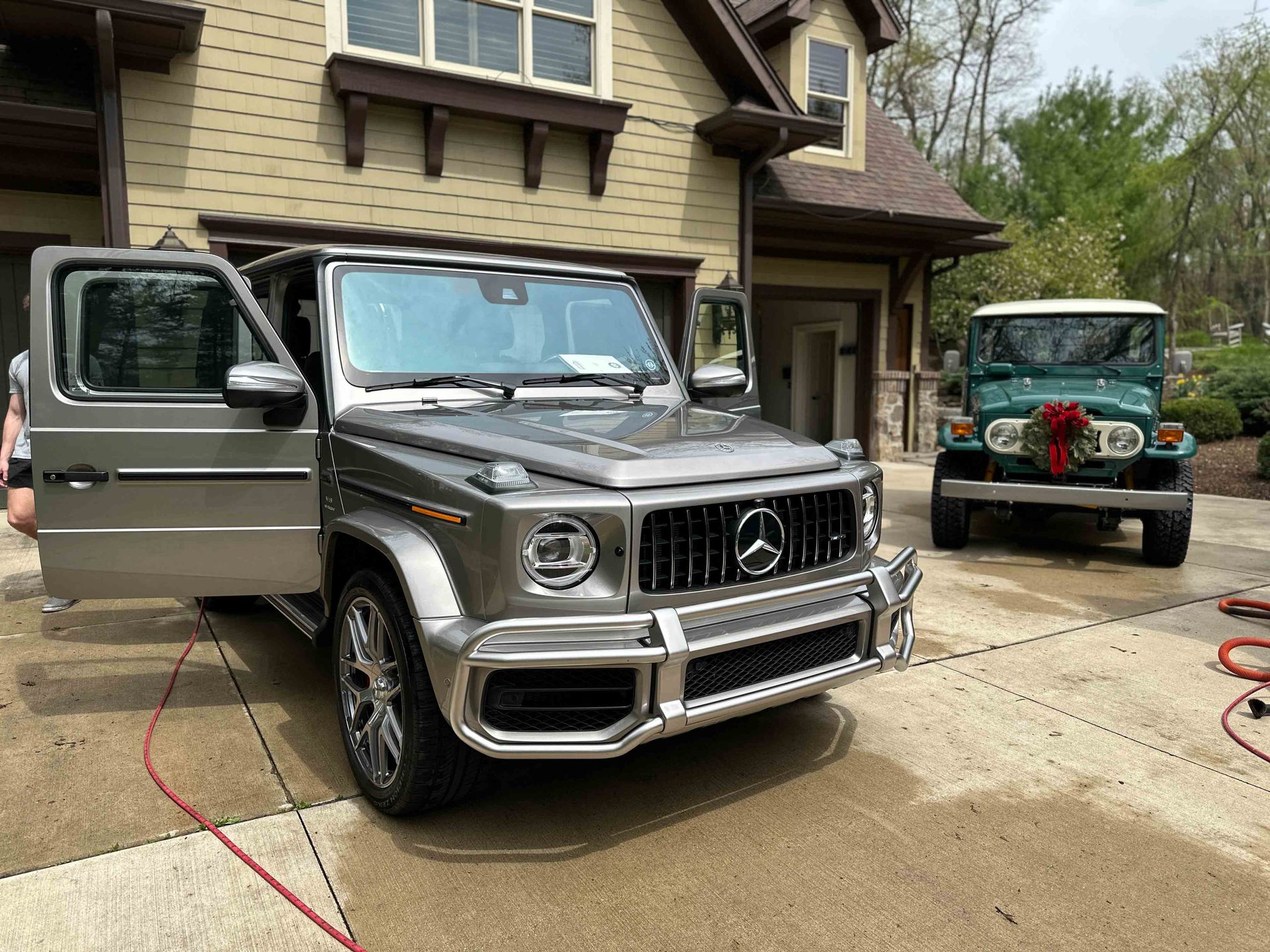 Silver Mercedes-Benz G-Class SUV parked next to a teal vintage Jeep in front of a house with a beige exterior.