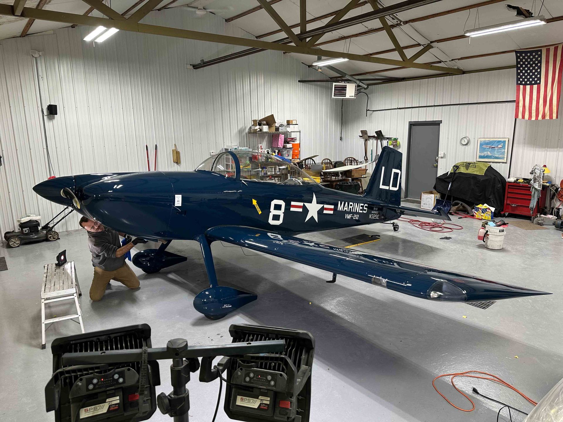 Person working on dark blue airplane in a hangar, with American flag on wall.