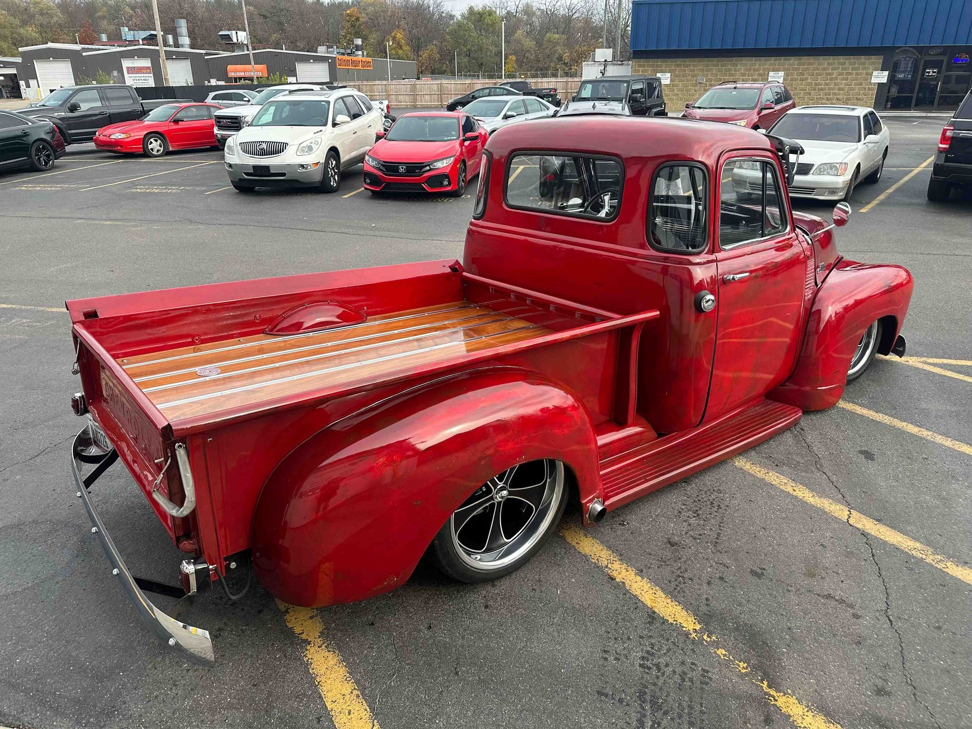 Red classic pickup truck parked in a lot, lowered with wood bed, other vehicles in background.
