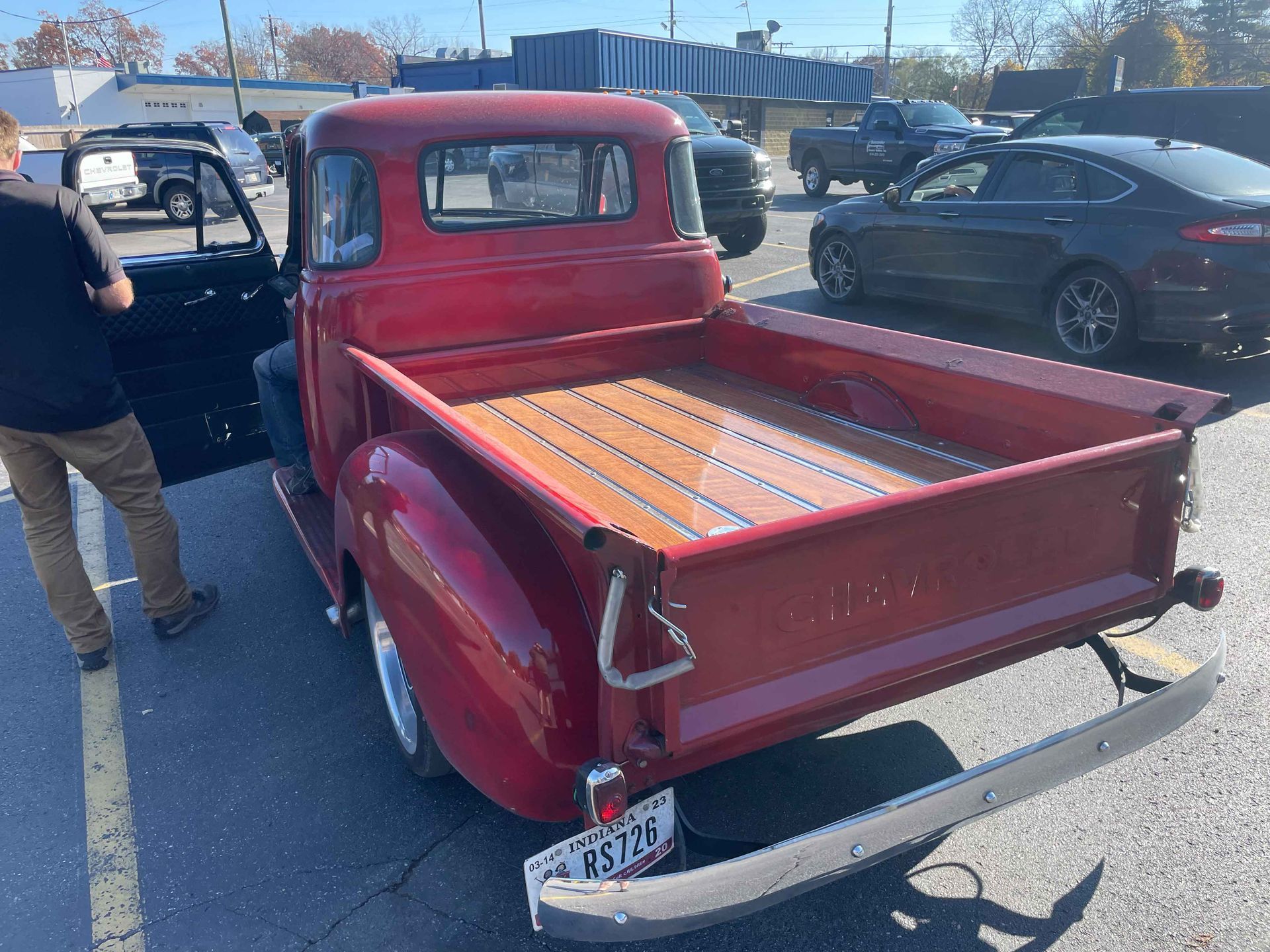 Red vintage pickup truck with wooden bed, parked in lot; person standing nearby.