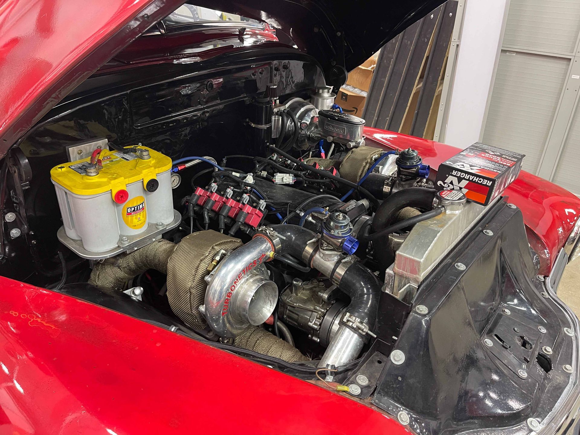 Red classic car engine bay with a large turbocharger and yellow battery.