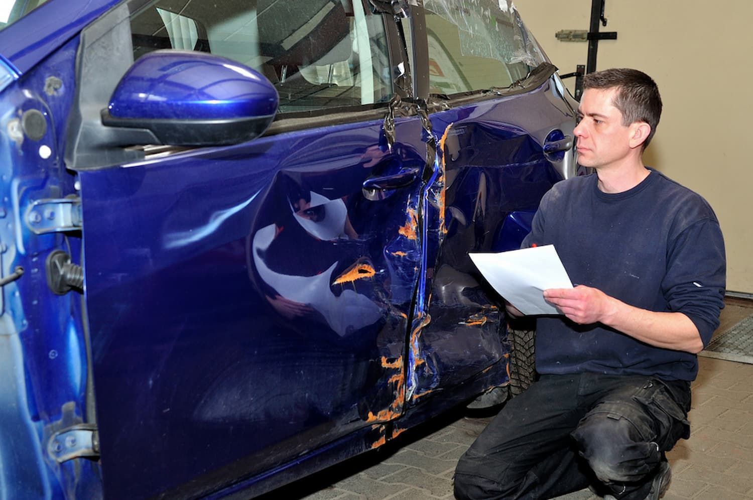 A Man Is Kneeling in Front of A Damaged Blue Car — Chinderah Smash Repairs in Chinderah, NSW