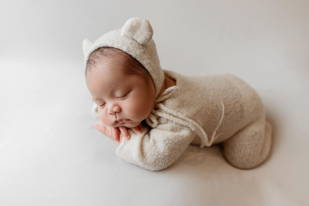 Newborn baby in beige bear outfit, sleeping on a white surface.