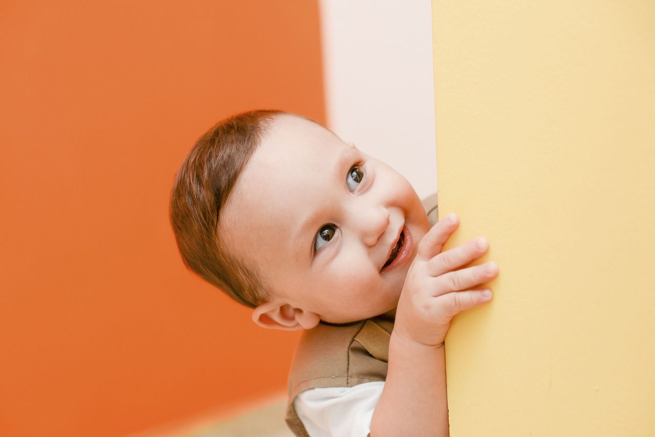 Smiling baby peeks from behind a yellow wall, orange background.