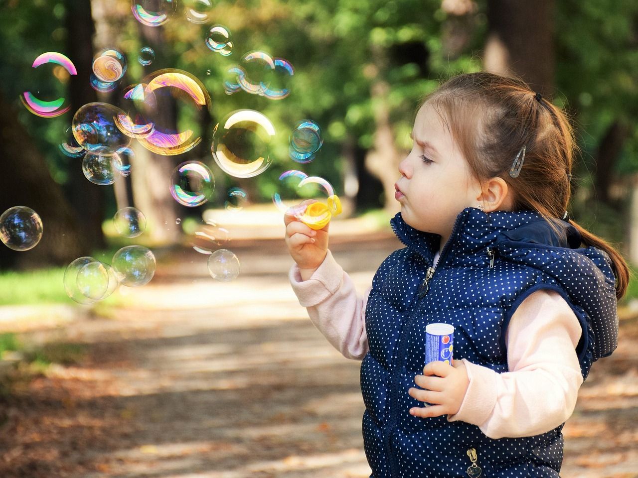 Girl blowing bubbles in a park, wearing a blue vest and a light pink shirt, sunny day.