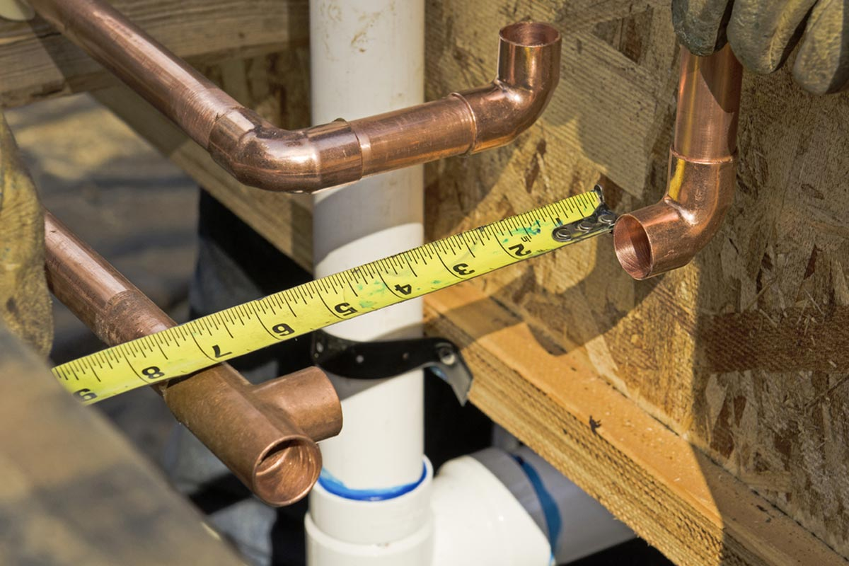 A person measures copper pipes with a tape measure near a white pipe and wooden structure.